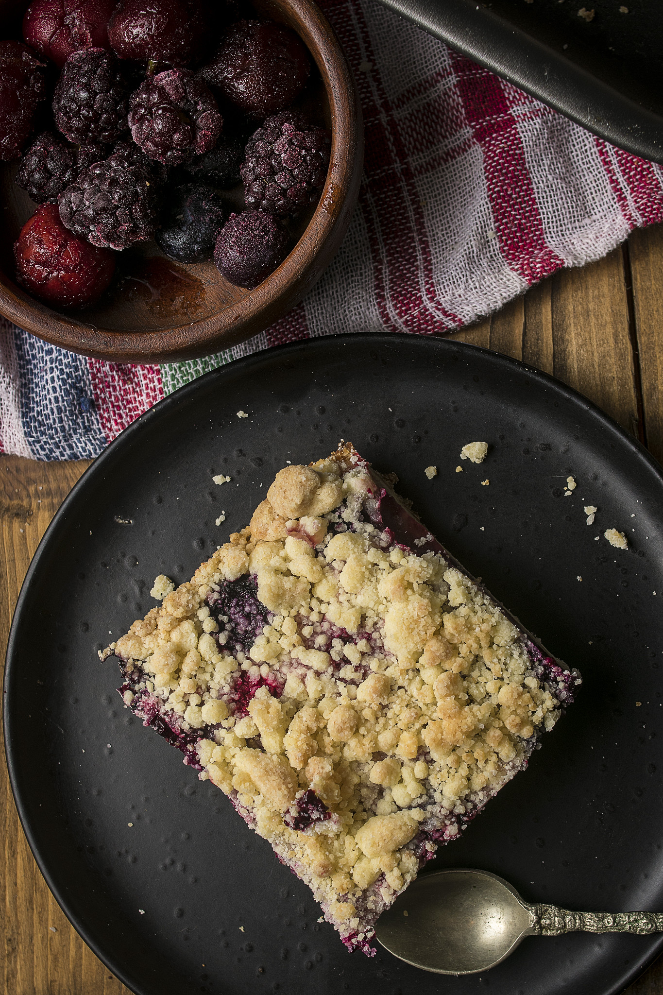 Overhead view of a slice of rustic Italian berry crumble tart on a black plate, with golden crumb topping, dark berry filling and frozen berries nearby.